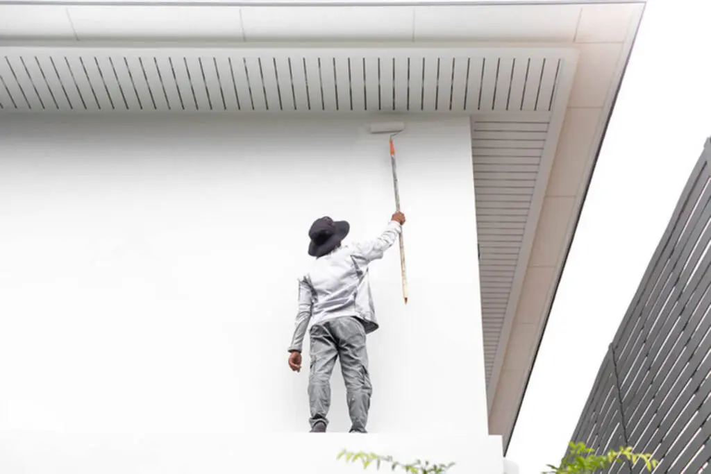 A professional painter using a roller on an exterior house wall during an outdoor painting project.