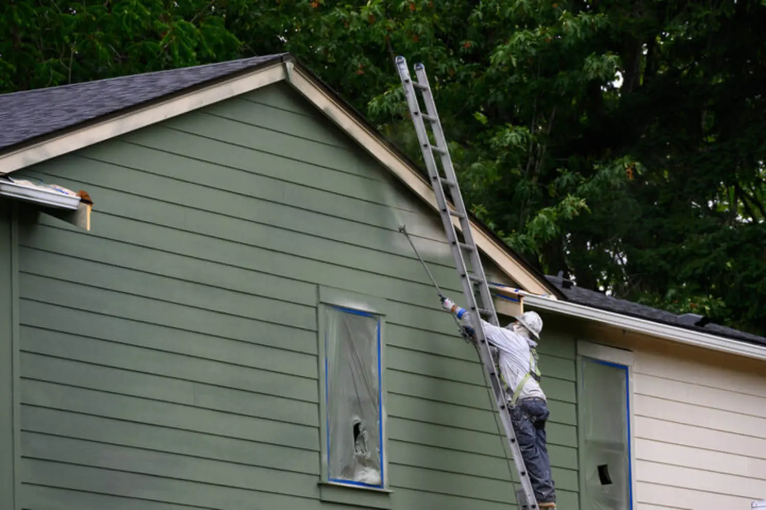 A professional painter on an extension ladder spraying exterior siding on a residential home with windows masked for protection.